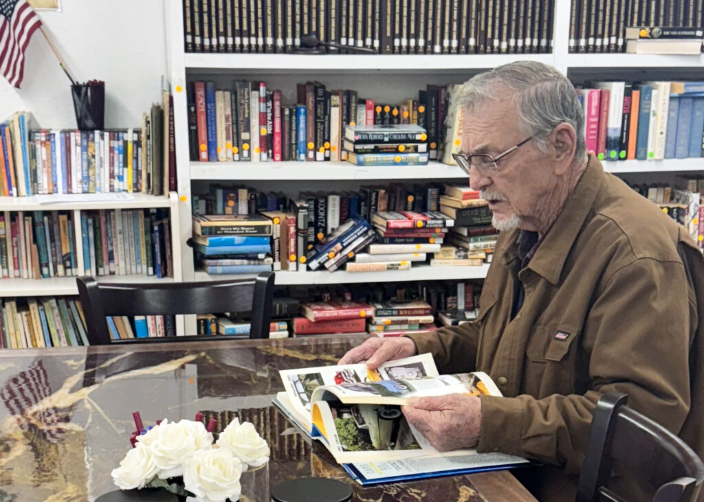 Photo of a library seated at a table looking through a book. Behind him are full bookshelves.